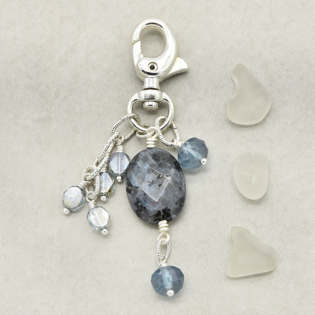 A silver-toned lobster clasp keychain charm featuring a large oval dark gray stone and three dangling bead strands with faceted blue-gray and iridescent beads, displayed on a light beige textured background with three small sea glass pieces to the right.