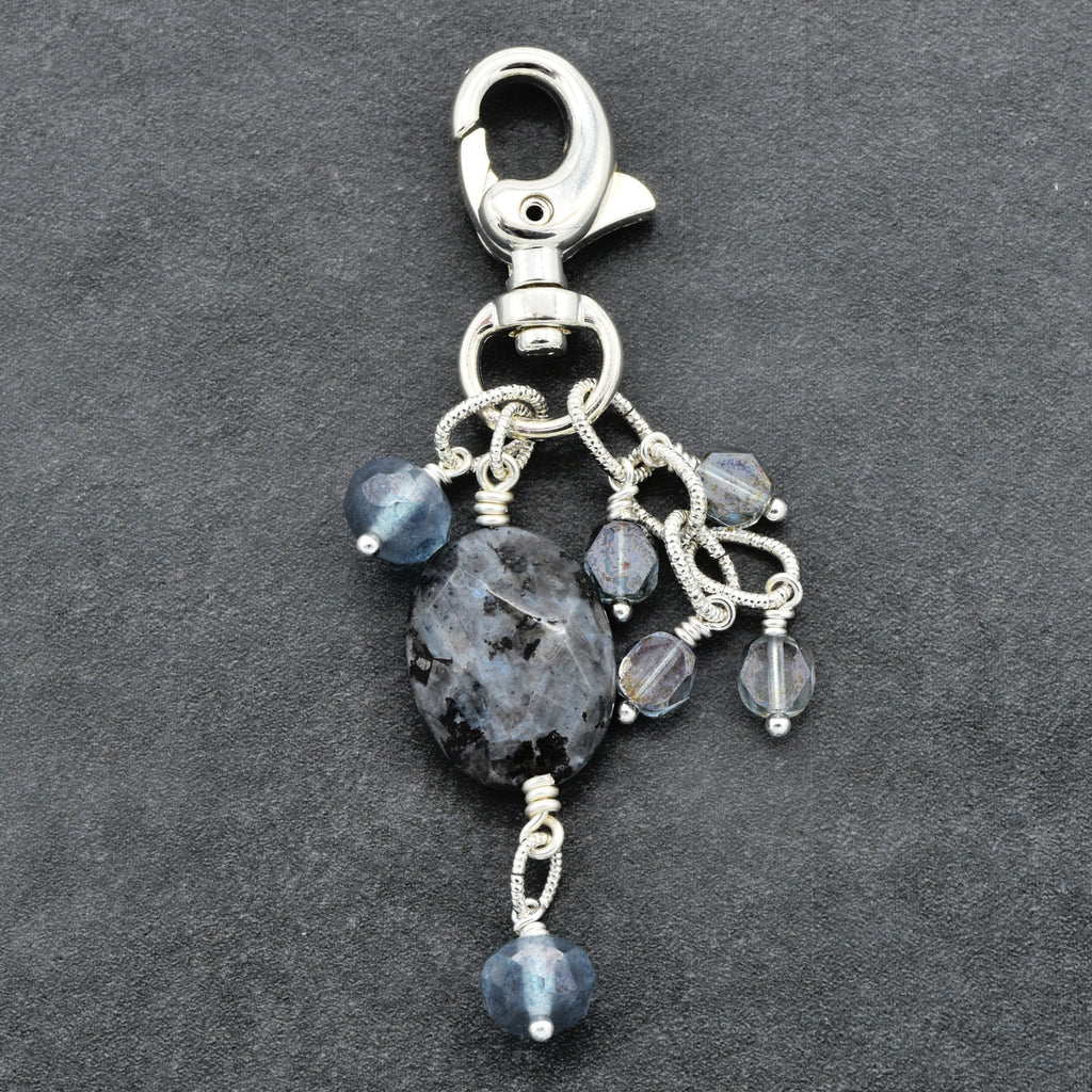A silver-toned lobster clasp keychain charm featuring a large dark gray agate stone and several smaller faceted blue-gray beads, displayed on a dark textured background.