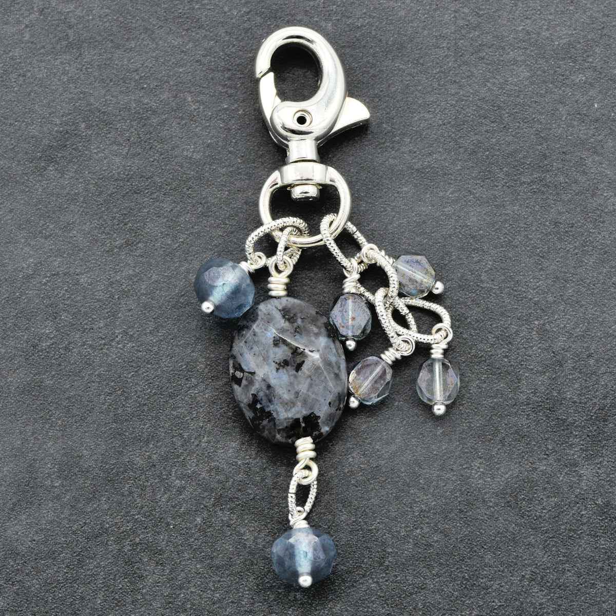A silver-toned lobster clasp keychain charm featuring a large dark gray agate stone and several smaller faceted blue-gray beads, displayed on a dark textured background.