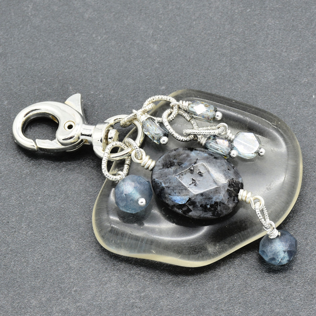 A silver-toned lobster clasp keychain or bag charm featuring a clear, irregularly shaped stone base with dangling blue-gray beads, a faceted dark stone, and clear crystal-like beads on a dark gray textured background.