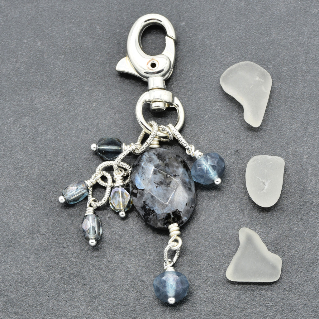 A handmade silver-toned keychain charm featuring a large faceted dark gray stone, blue-gray beads, and small iridescent beads, displayed next to three pieces of natural sea glass on a dark gray textured background.