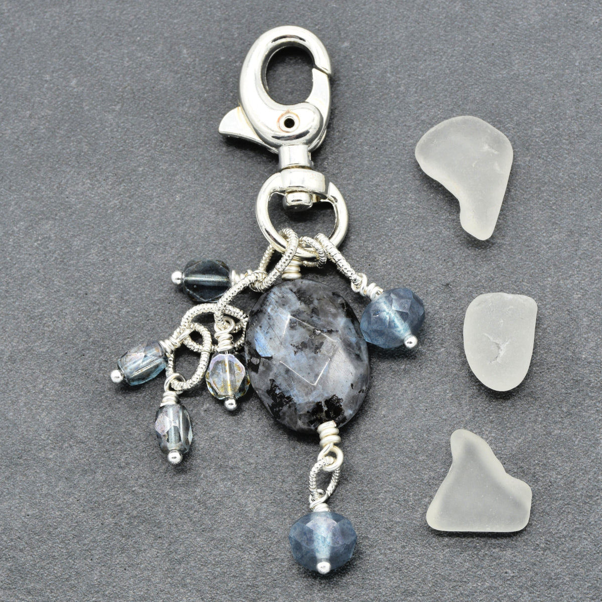 A handmade silver-toned keychain charm featuring a large faceted dark gray stone, blue-gray beads, and small iridescent beads, displayed next to three pieces of natural sea glass on a dark gray textured background.