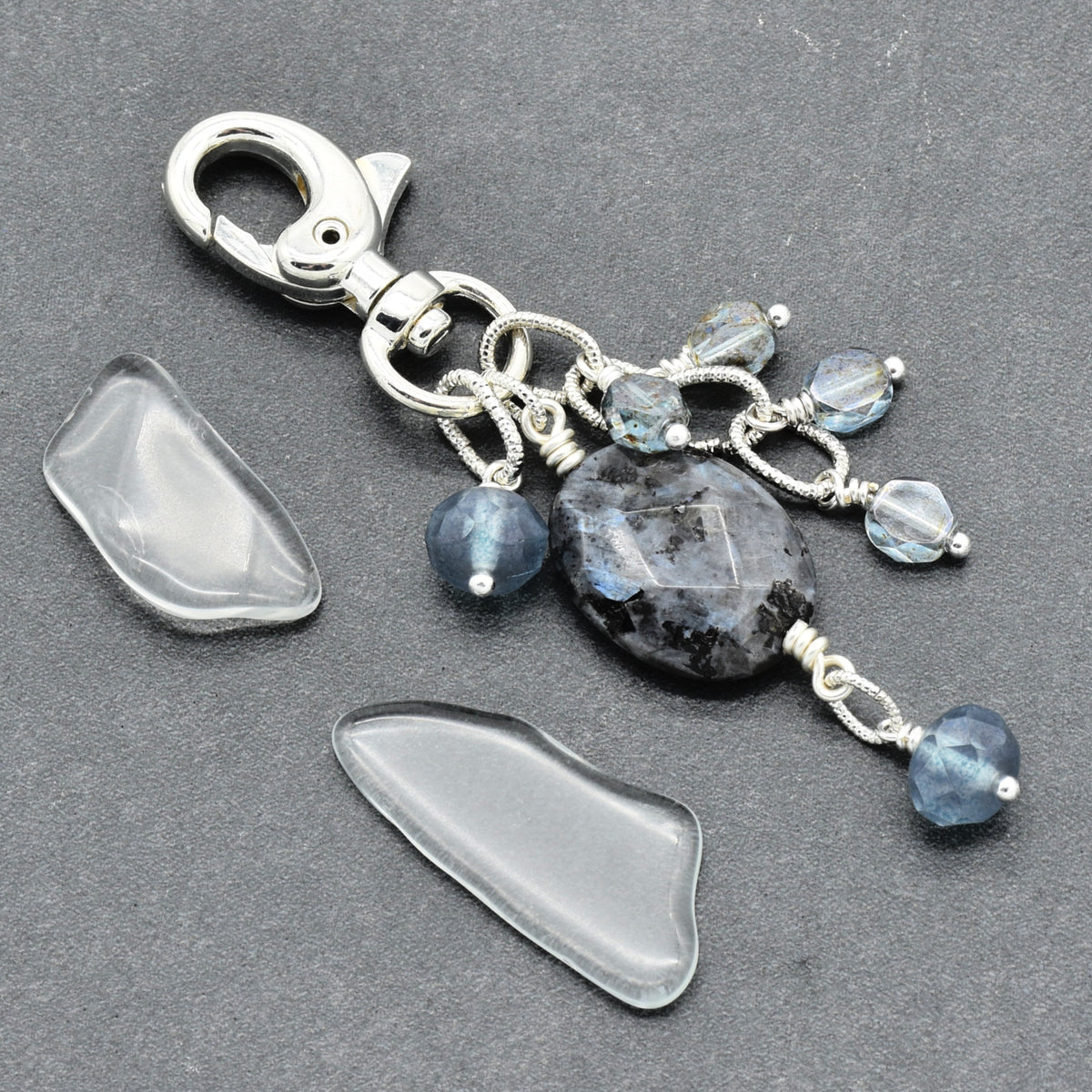 A handmade silver keychain featuring a large dark blue feldspar stone, smaller faceted blue beads, and two clear, polished raw stone pieces placed beside it on a dark gray textured background.
