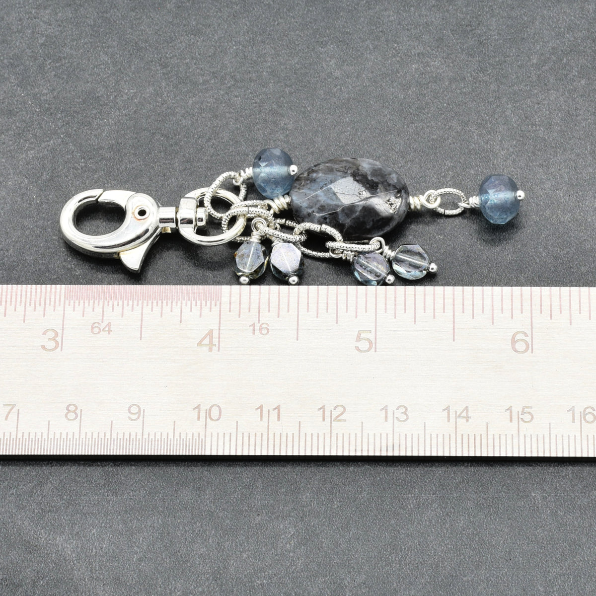 A silver-toned metal keychain or bag charm with faceted blue beads and clear crystal drops, shown above a ruler on a dark gray textured background.