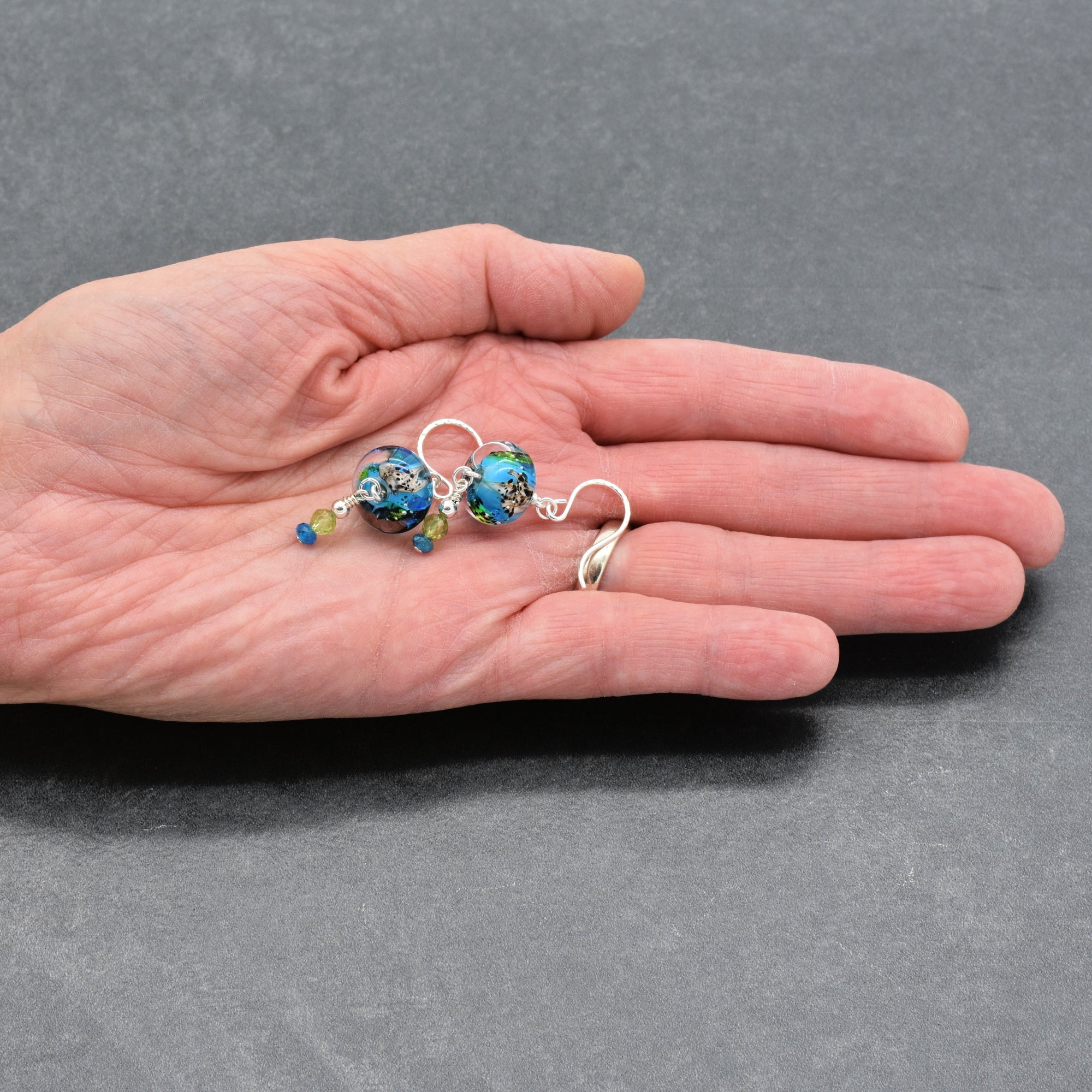 Artist's hand holding colorful artisan lampwork glass earrings with sterling silver ear wires, peridot and apatite on a gray surface - handmade with love by Ruby Acorn