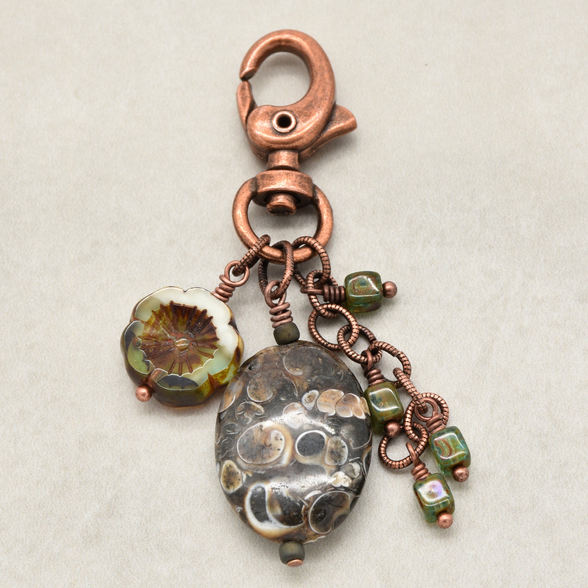 A handmade copper keychain featuring a large oval agate bead with a spotted pattern, a smaller round patterned bead, and green glass beads wrapped in copper wire, shown on a neutral light gray background - made with love in Colorado by Ruby Acorn.