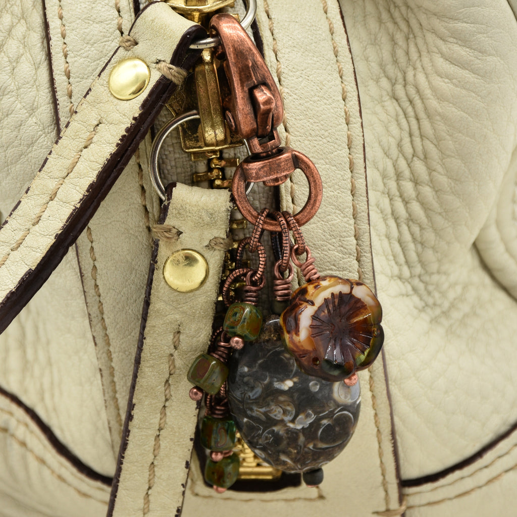 Close-up of a handbag hardware charm with copper-toned metal rings, a large dark marbled bead, a floral glass bead, and small green beads attached to a cream-colored leather strap - made with love in Colorado by Ruby Acorn.