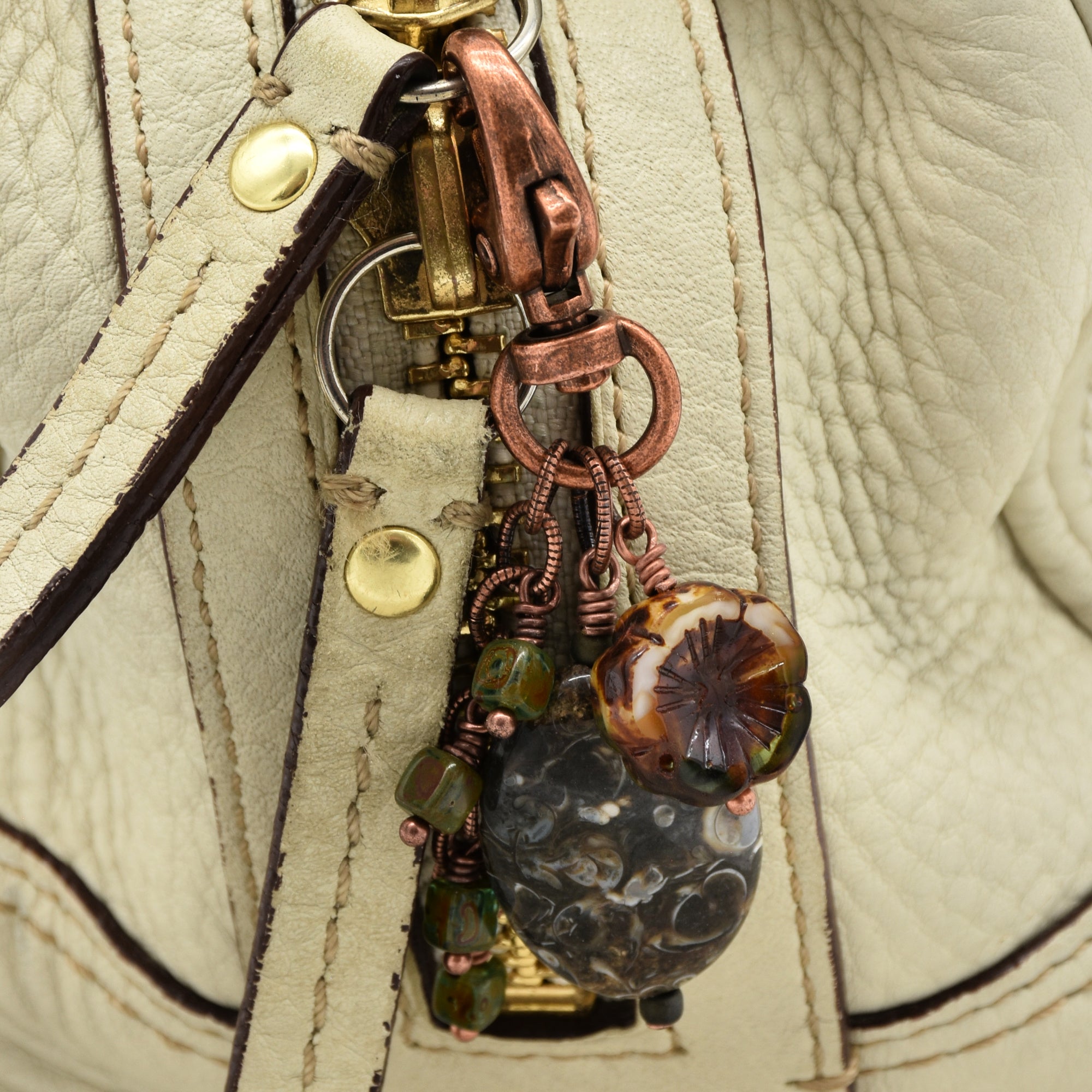 Close-up of a handbag hardware charm with copper-toned metal rings, a large dark marbled bead, a floral glass bead, and small green beads attached to a cream-colored leather strap - made with love in Colorado by Ruby Acorn.