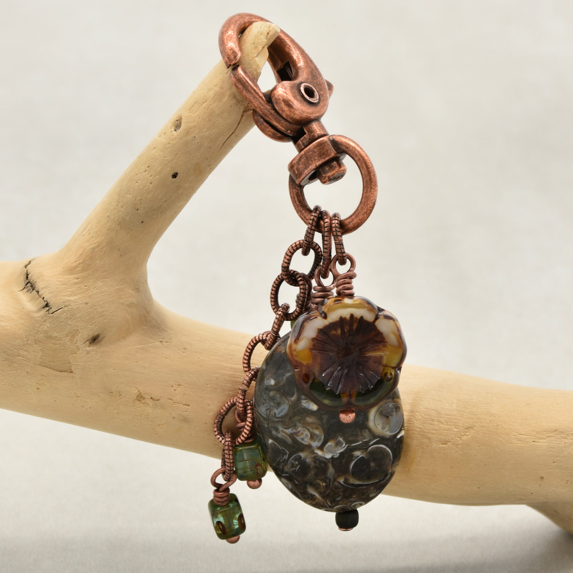A handmade copper keychain with a large textured dark bead, green glass beads, and a floral glass charm, hanging from a copper lobster clasp on a light wood branch - made with love in Colorado by Ruby Acorn.