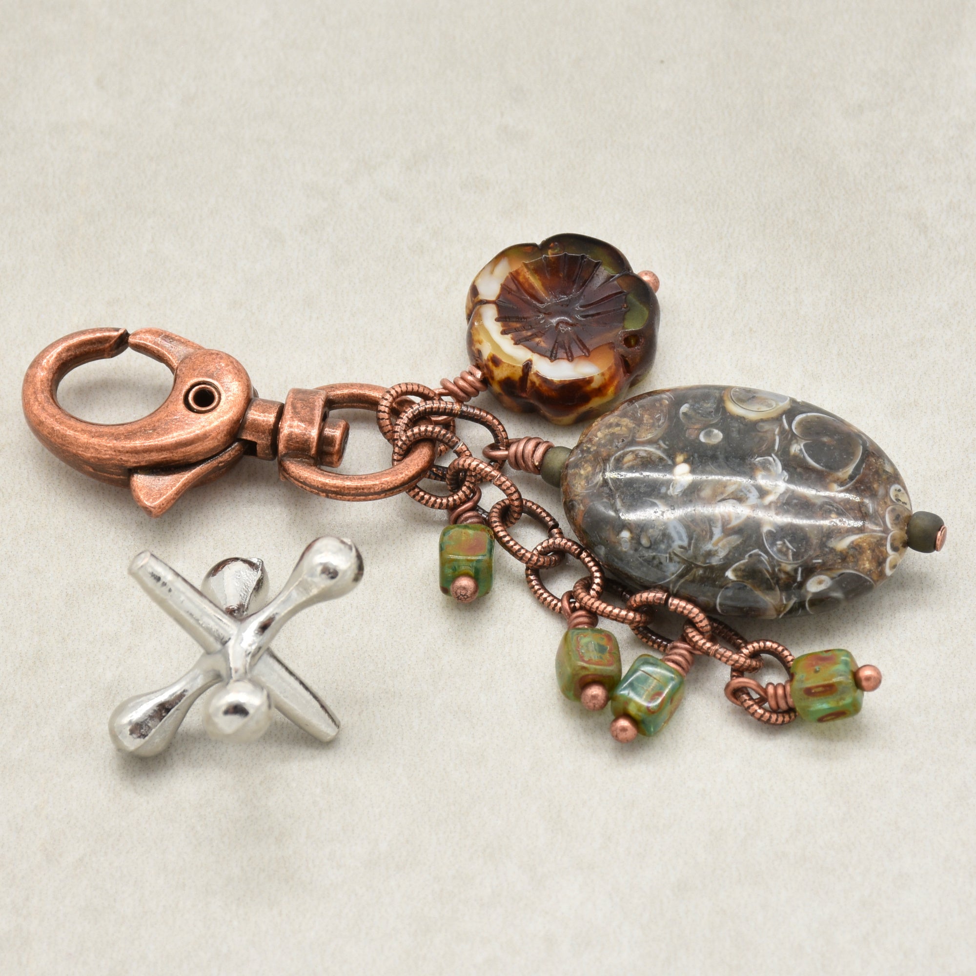 A handmade keychain featuring a copper clasp, a large marbled agate bead, a copper-wrapped chain, small green beads, and a flower-shaped amber-brown bead, displayed on a neutral light gray background - made with love in Colorado by Ruby Acorn.