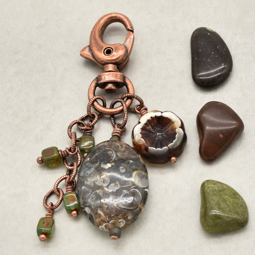 A handmade copper wire jewelry charm featuring a large agate bead, a flower bead, and green stone beads, displayed next to three polished stones on a neutral background - made with love in Colorado by Ruby Acorn.