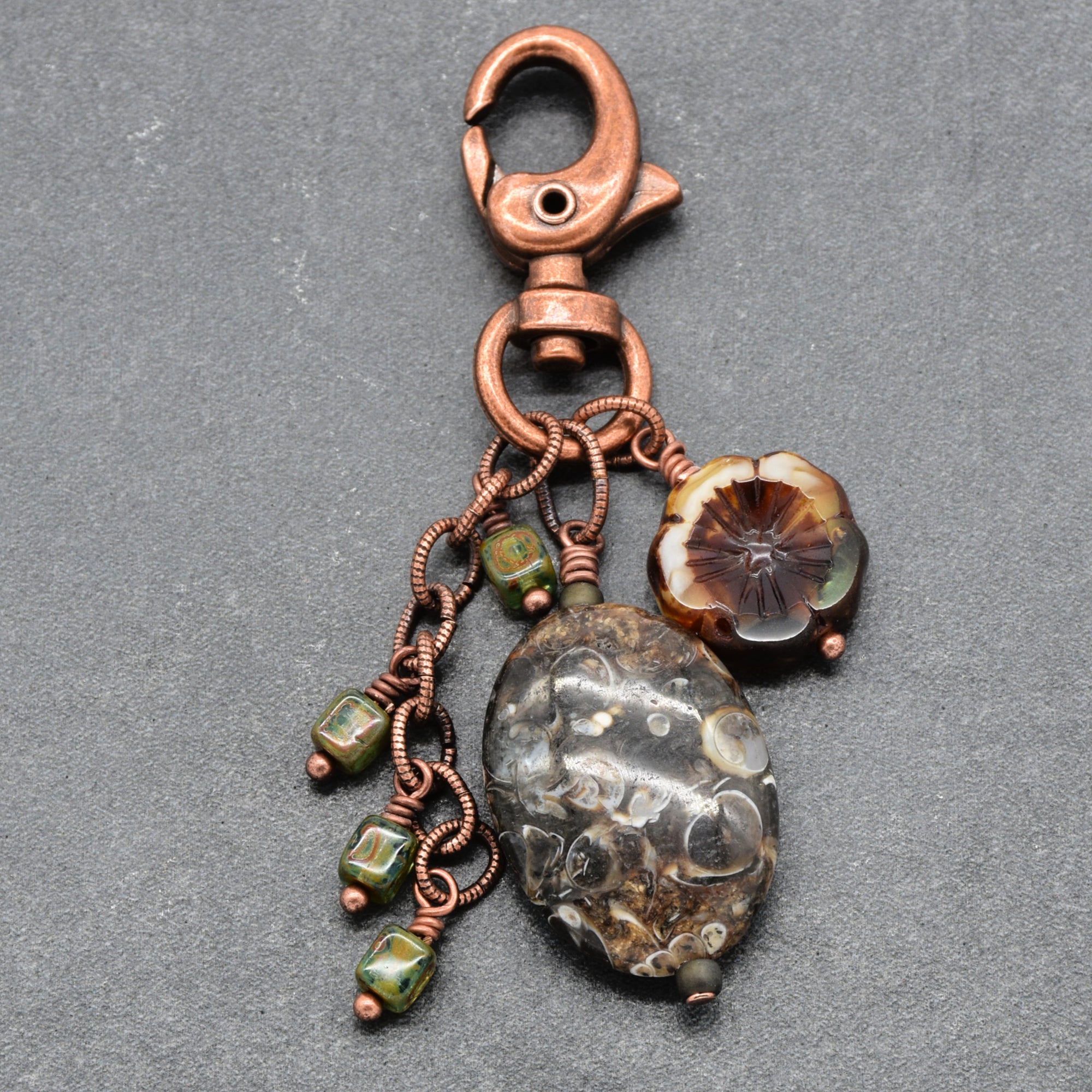 A copper wire-wrapped keychain or bag charm with a lobster clasp, featuring a large polished agate stone, a small flower bead, and three green beads on a textured gray background - made with love in Colorado by Ruby Acorn.