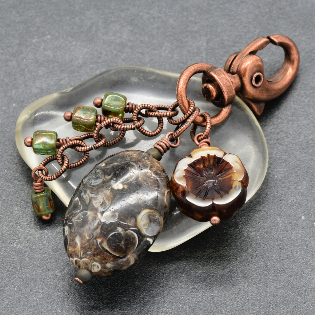 A handmade jewelry charm or keychain featuring a large textured dark brown stone bead, a carved floral bead with brown and cream layers, green beads, and copper wire chains attached to a copper clasp, all resting on a translucent glass dish over a dark gray background - made with love in Colorado by Ruby Acorn.