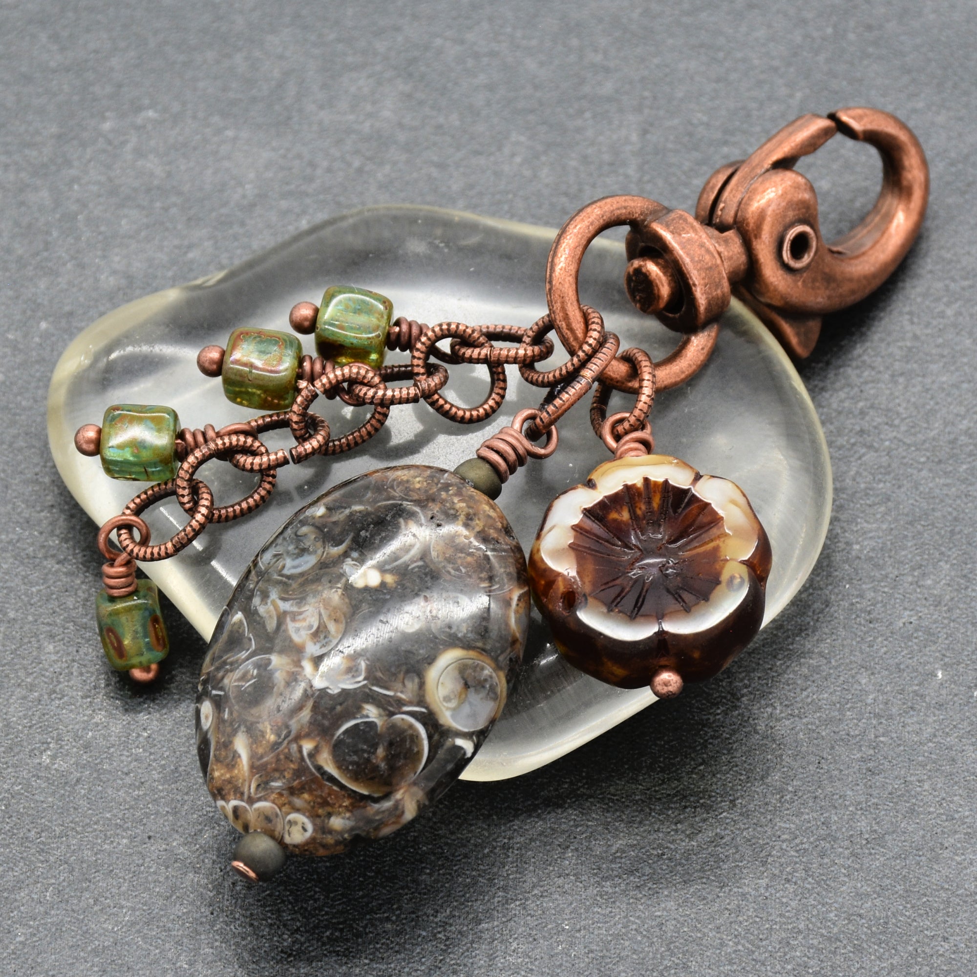 A handmade jewelry charm or keychain featuring a large textured dark brown stone bead, a carved floral bead with brown and cream layers, green beads, and copper wire chains attached to a copper clasp, all resting on a translucent glass dish over a dark gray background - made with love in Colorado by Ruby Acorn.