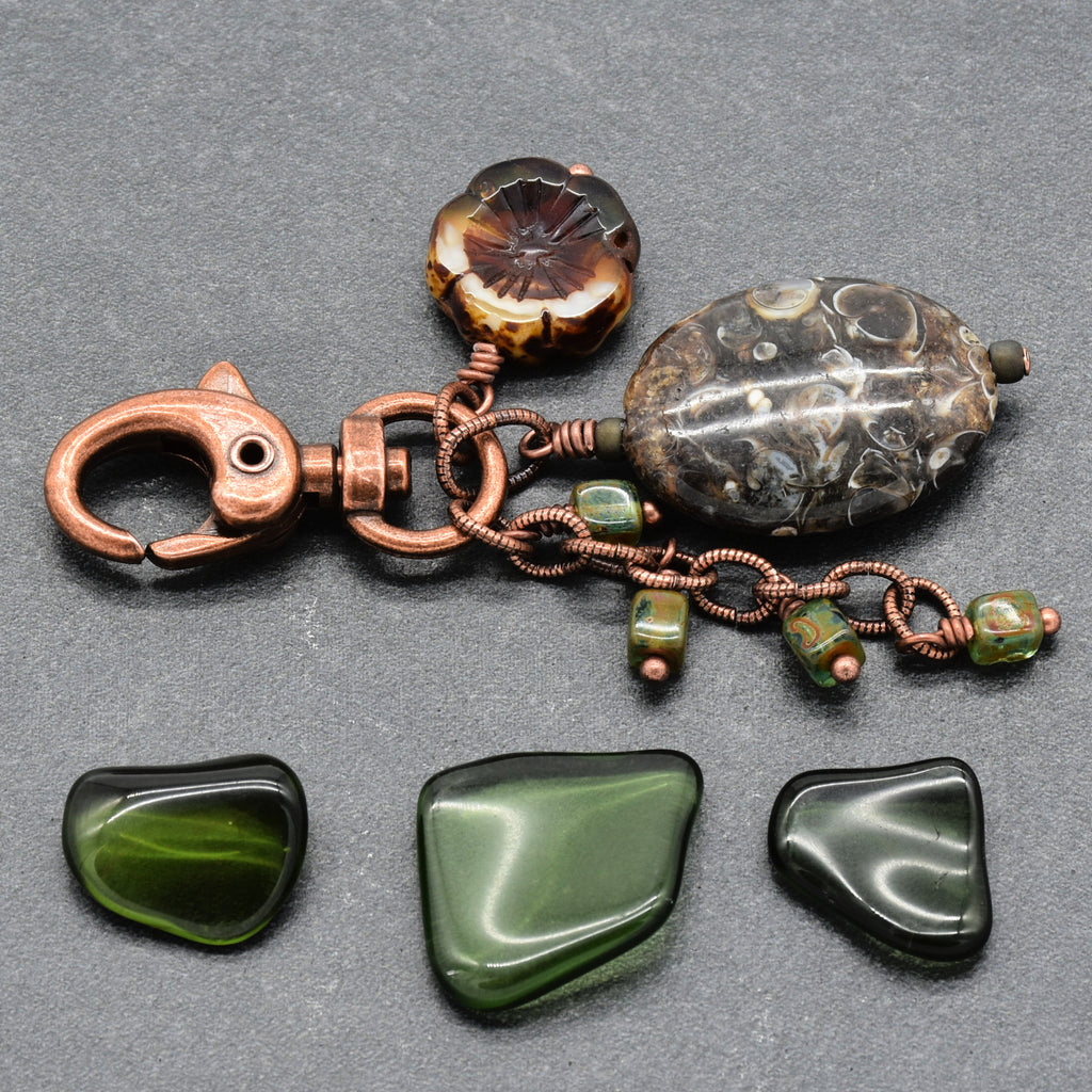A handmade copper-wrapped keychain or bag charm with a large polished dark stone bead, a flower-shaped amber-brown bead, and three green stone chips, displayed on a dark gray background - made with love in Colorado by Ruby Acorn.