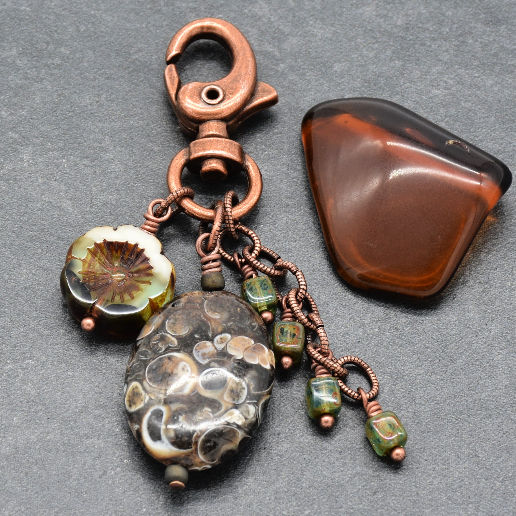 A handmade keychain or bag charm featuring a copper-toned lobster clasp, a large patterned brown and cream stone bead, a smaller green beaded charm, and a polished brown stone piece, displayed on a dark gray textured background - made with love in Colorado by Ruby Acorn.