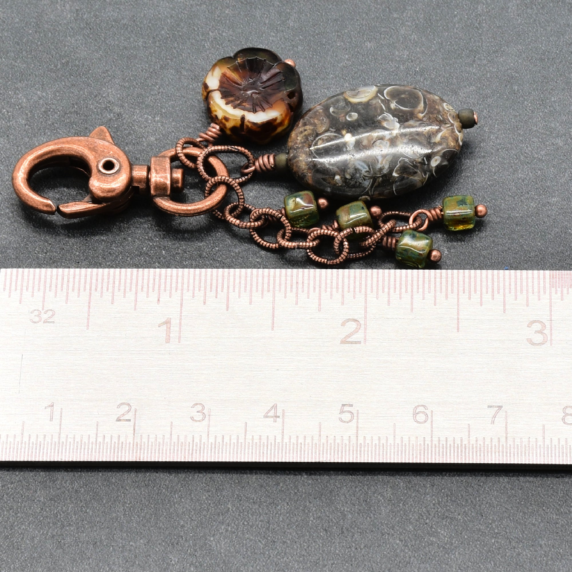 A handmade copper and glass keychain charm with a large polished agate stone, a flower-shaped glass bead, and small green glass beads, shown next to a metal ruler for scale on a dark gray background - made with love in Colorado by Ruby Acorn.