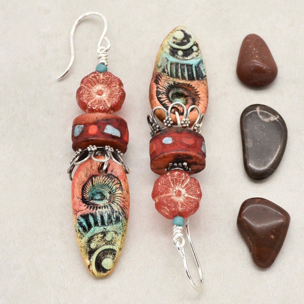 A pair of handmade dangle earrings with silver-tone fishhook wires, featuring red and orange patterned beads, turquoise accents, and decorative silver spacers, displayed next to four smooth brown stones on a light beige background - made with love in Colorado by Ruby Acorn.