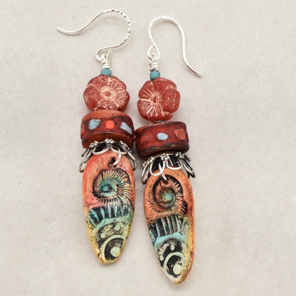 A pair of handmade dangle earrings featuring textured red and orange beads, turquoise accents, and silver-tone hooks on a neutral beige background - made with love in Colorado by Ruby Acorn.