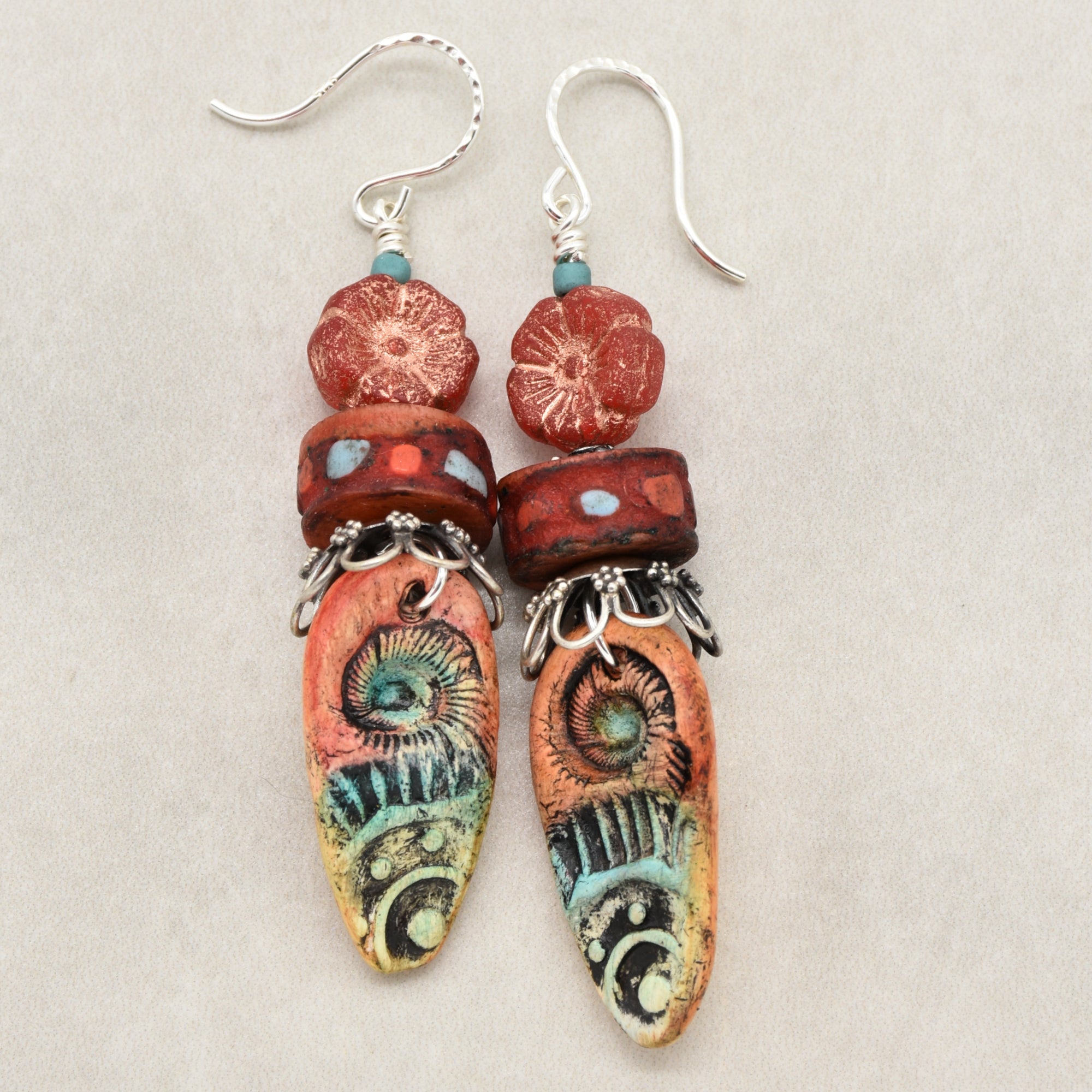 A pair of handmade dangle earrings featuring textured red and orange beads, turquoise accents, and silver-tone hooks on a neutral beige background - made with love in Colorado by Ruby Acorn.