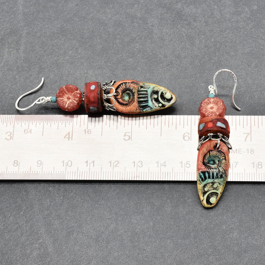 A pair of handmade beaded earrings displayed on a gray background with a metal ruler for scale. The earrings feature a silver fishhook ear wire, a small round red bead, a larger textured red bead, and a long, narrow central bead decorated with turquoise, green, and orange patterns - made with love in Colorado by Ruby Acorn.