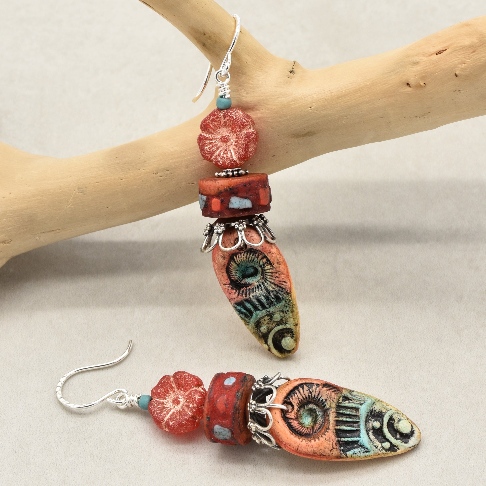 A pair of handmade dangle earrings displayed on a light wood branch and against a neutral beige background. The earrings feature a silver-tone fishhook, a small red flower bead, a red cylindrical bead with turquoise accents, and a large teardrop-shaped polymer clay charm decorated with a colorful tribal design in orange, green, and black - made with love in Colorado by Ruby Acorn.