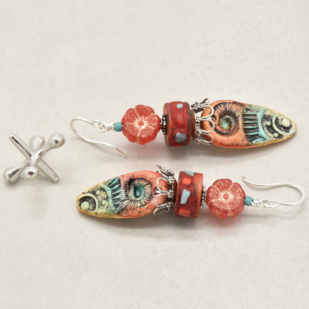 A pair of handmade dangle earrings with colorful, patterned marquise-shaped beads, red flower beads, and turquoise spacer beads on silver fishhook wires, shown alongside a jack on a light beige background - made with love in Colorado by Ruby Acorn.