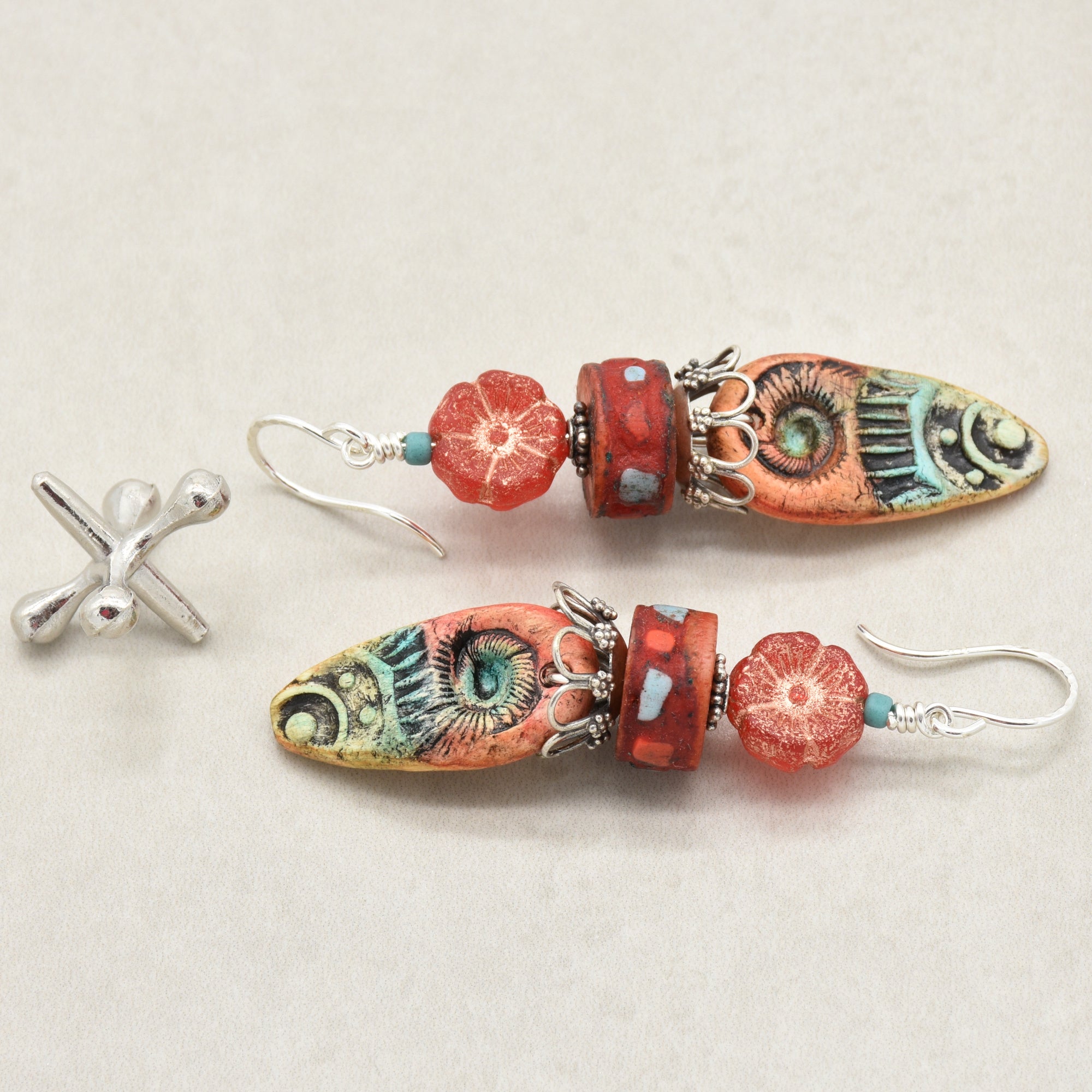 A pair of handmade dangle earrings with colorful, patterned marquise-shaped beads, red flower beads, and turquoise spacer beads on silver fishhook wires, shown alongside a jack on a light beige background - made with love in Colorado by Ruby Acorn.