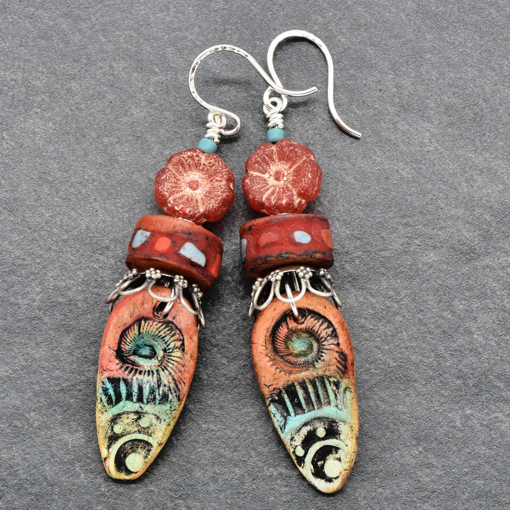 A pair of handmade dangle earrings with silver fishhook wires, turquoise beads, red patterned beads, and long oval charms decorated with colorful spiral and swirl motifs in orange, turquoise, black, and cream, displayed on a textured gray background - made with love in Colorado by Ruby Acorn.