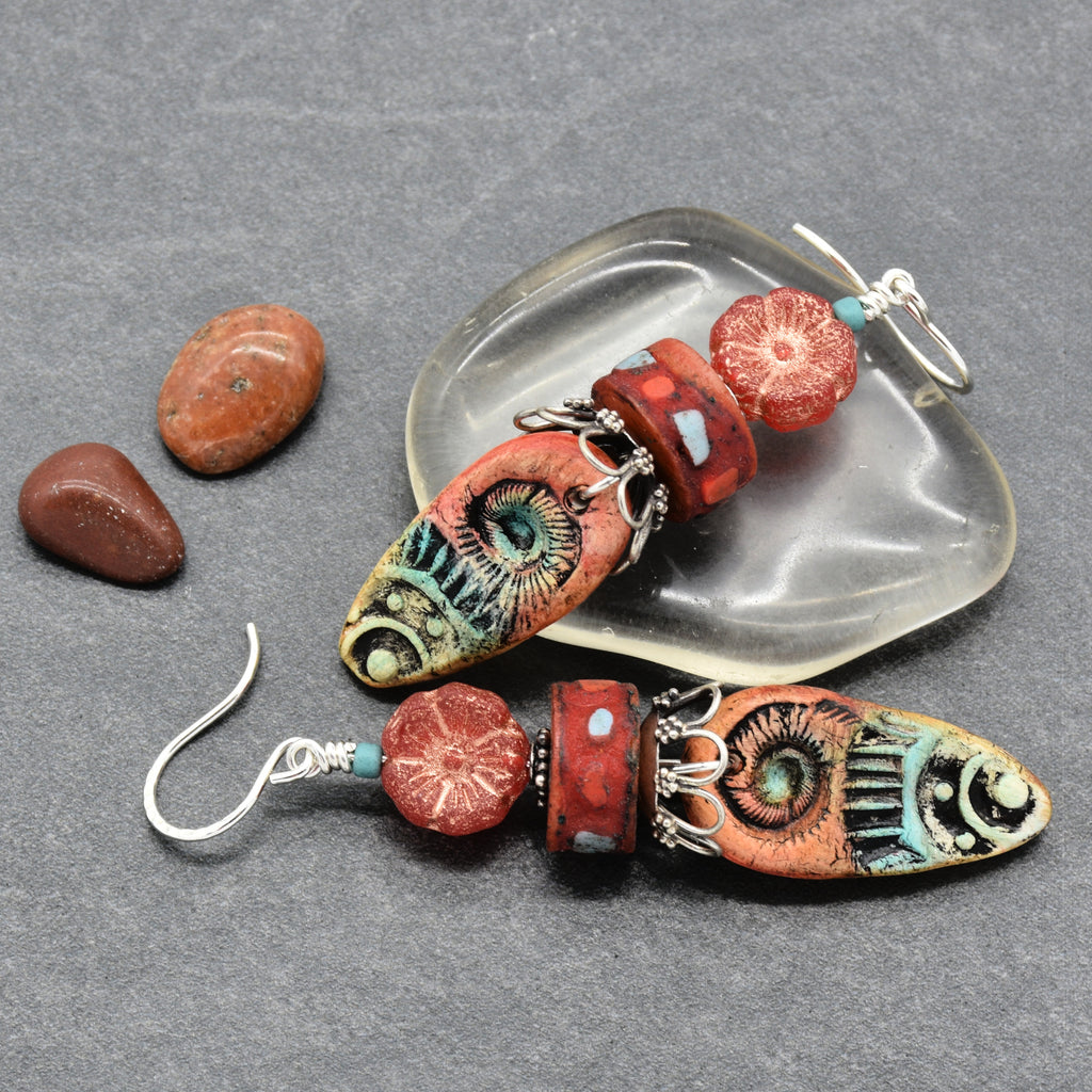 A pair of handmade dangle earrings with colorful patterned beads and silver hooks, displayed on a gray textured background with a clear dish and small stones - made with love in Colorado by Ruby Acorn.