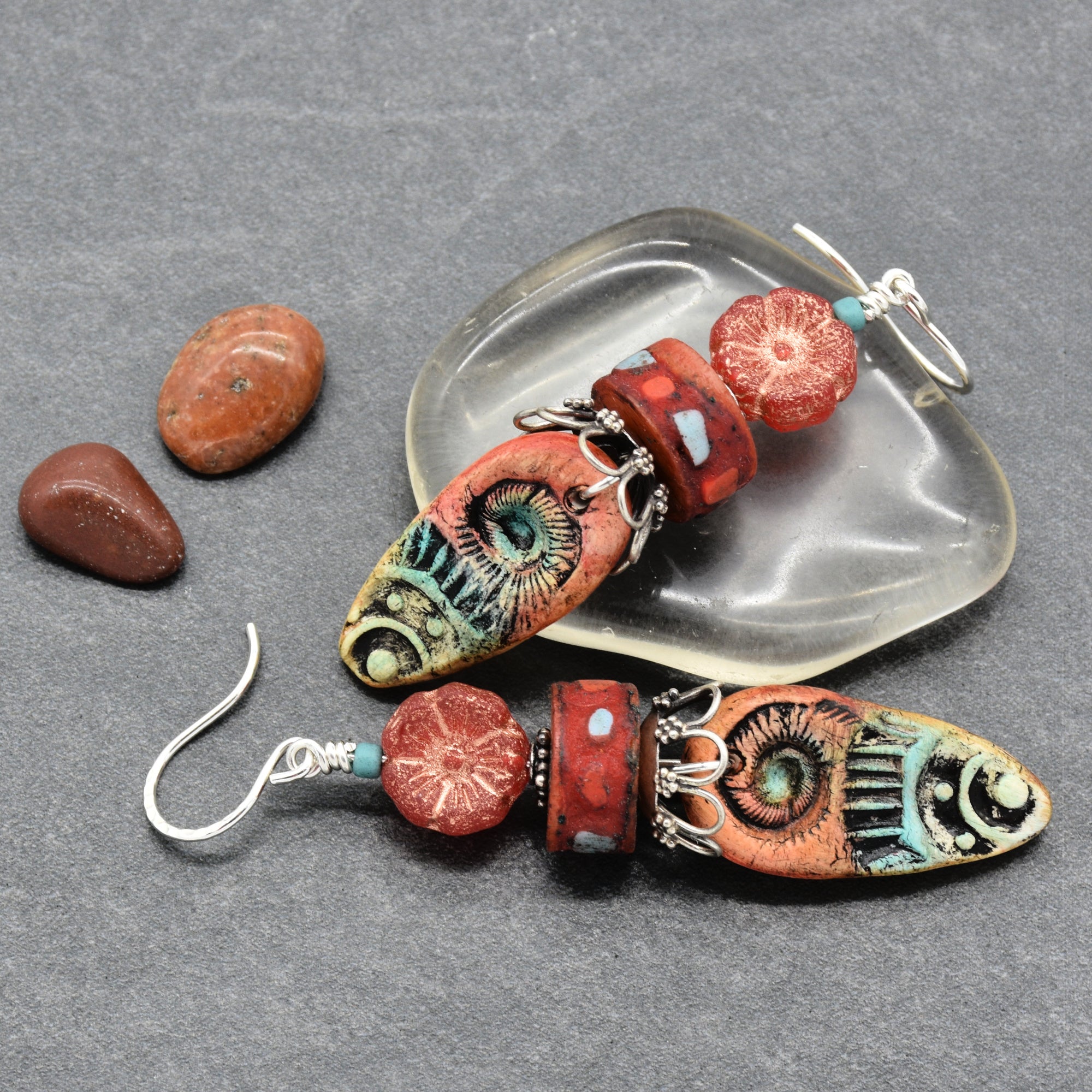 A pair of handmade dangle earrings with colorful patterned beads and silver hooks, displayed on a gray textured background with a clear dish and small stones - made with love in Colorado by Ruby Acorn.