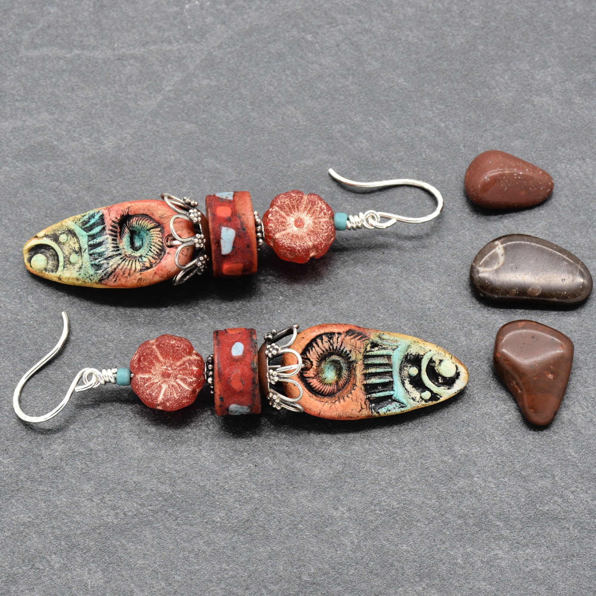 A pair of handmade dangle earrings with turquoise and red beads, displayed on a gray textured background next to four polished river stones - made with love in Colorado by Ruby Acorn.