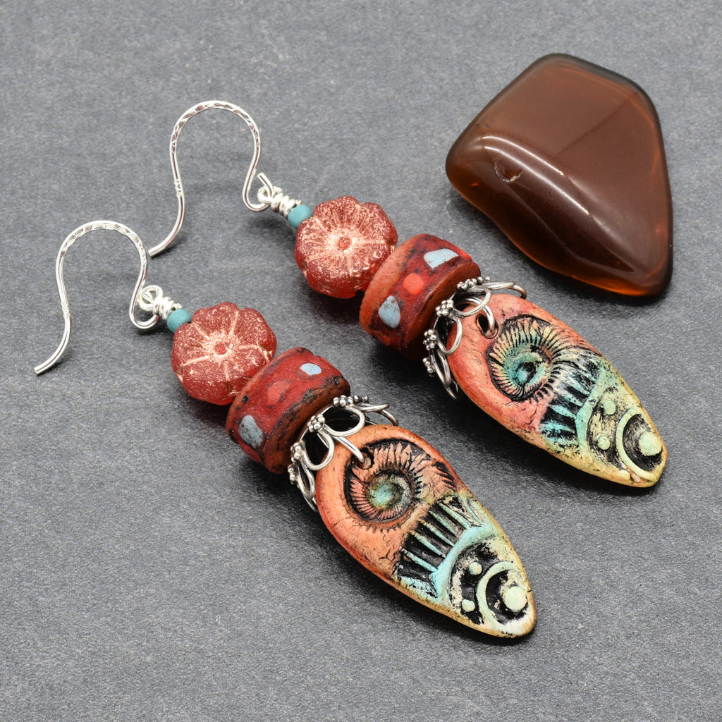 A pair of handmade dangle earrings with silver fishhook wires, turquoise accent beads, and terracotta flower beads, featuring long, hand-painted arrowhead-shaped drops with a stylized eye and tribal patterns in red, turquoise, black, and gold. A polished brown stone is displayed in the background on a textured gray surface - made with love in Colorado by Ruby Acorn. 