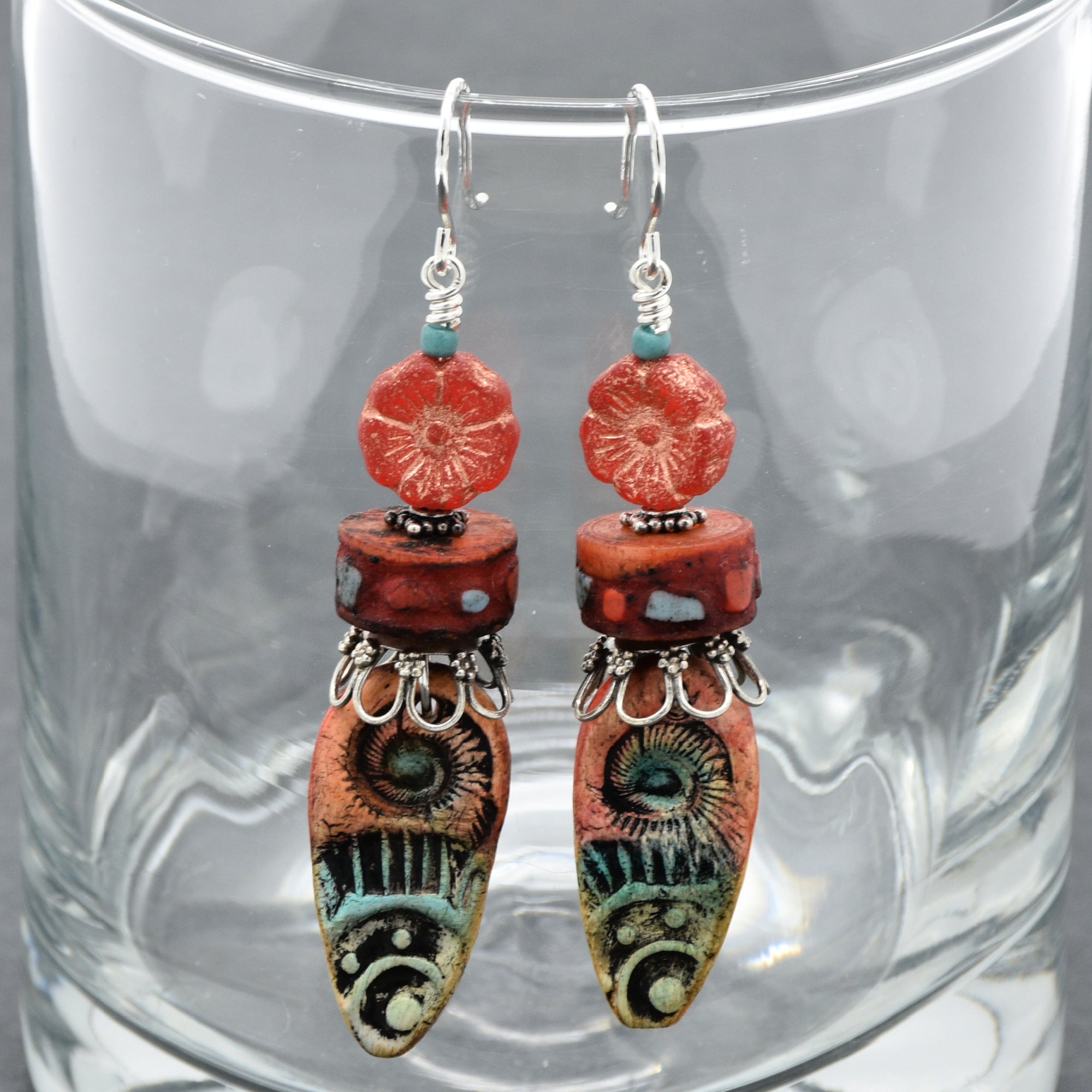 A pair of handmade dangle earrings with coral-red flower beads, patterned red and turquoise beads, and silver-toned metal details, hanging in front of a clear glass cup - made with love in Colorado by Ruby Acorn.