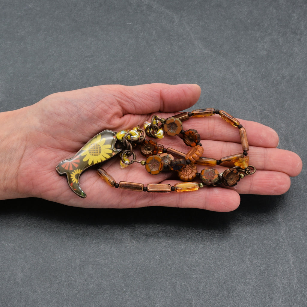 Hand holding a beaded necklace with a sunflower pendant on a gray background