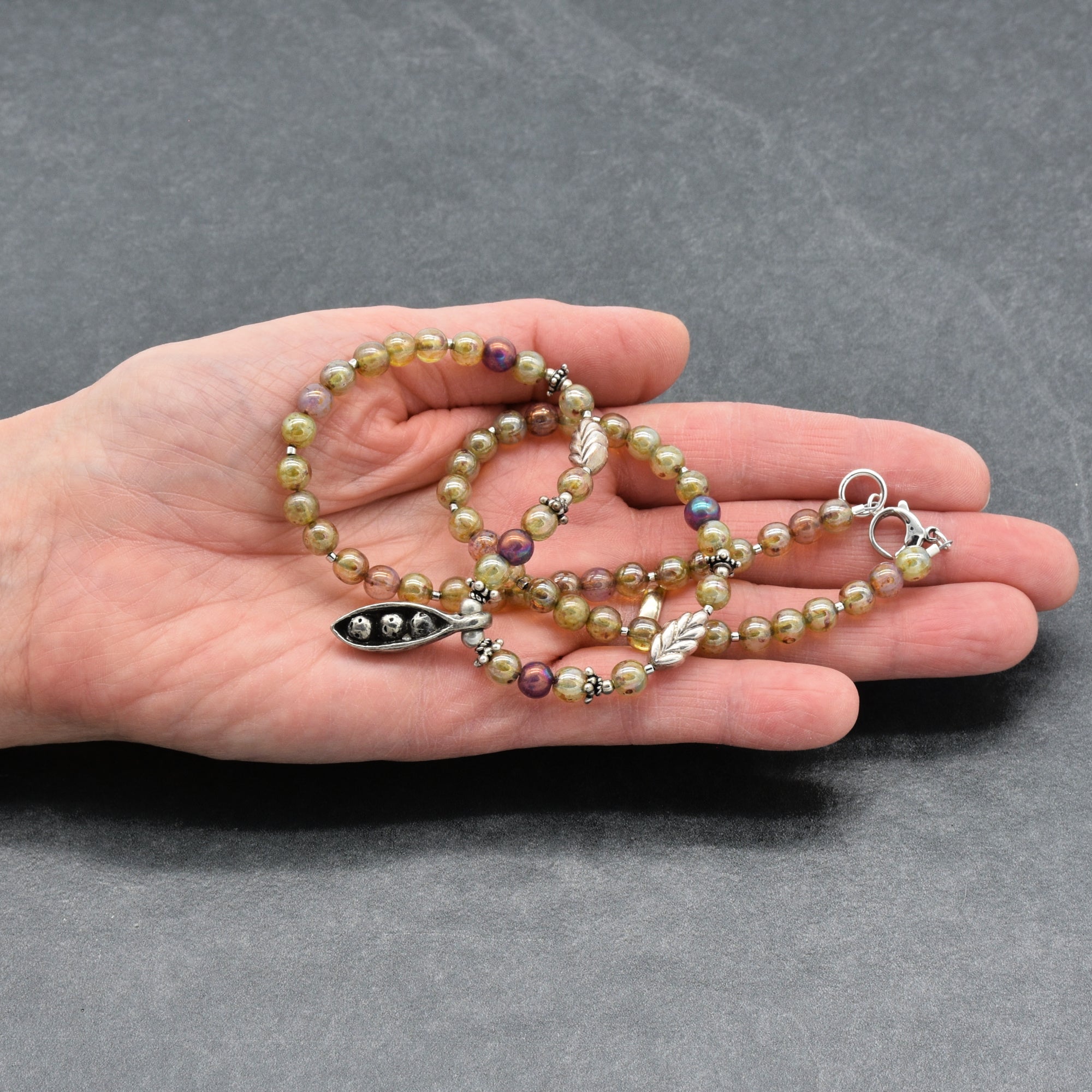 Beaded necklace with pewter peapod pendant held in a hand against a gray background