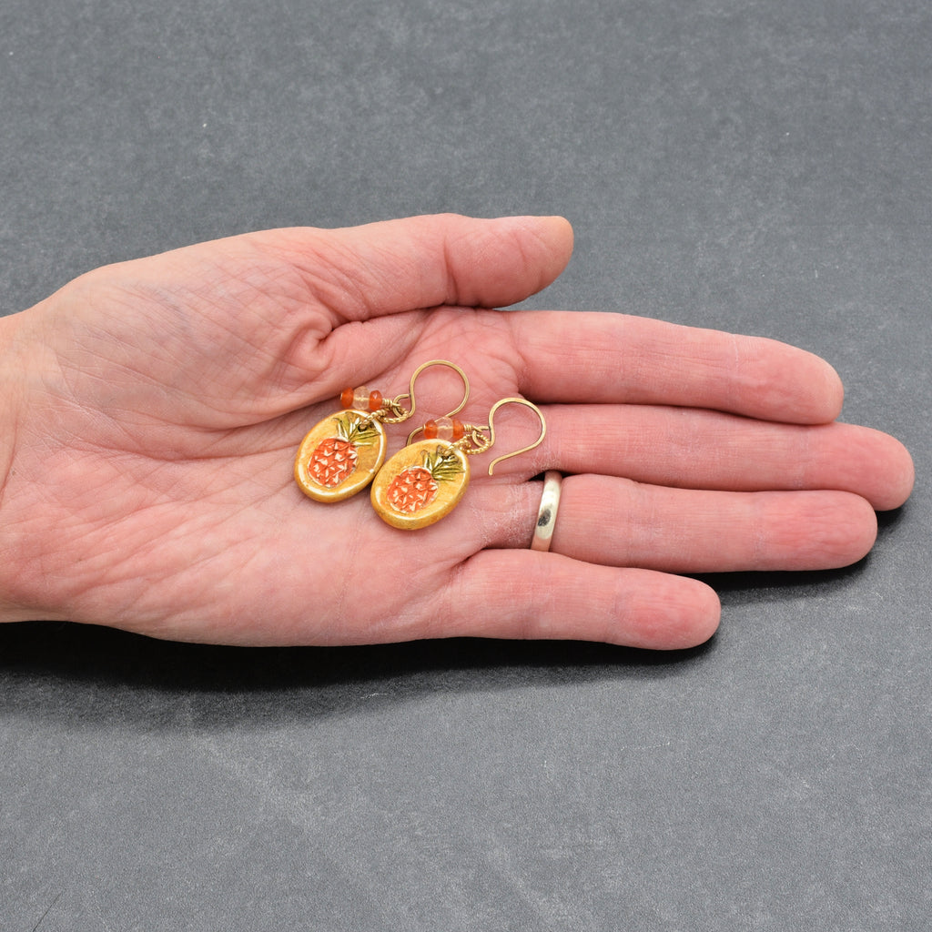 Artist's hand holding Pineapple charm earrings with brass ear wires on a gray surface - made with love by Ruby Acorn