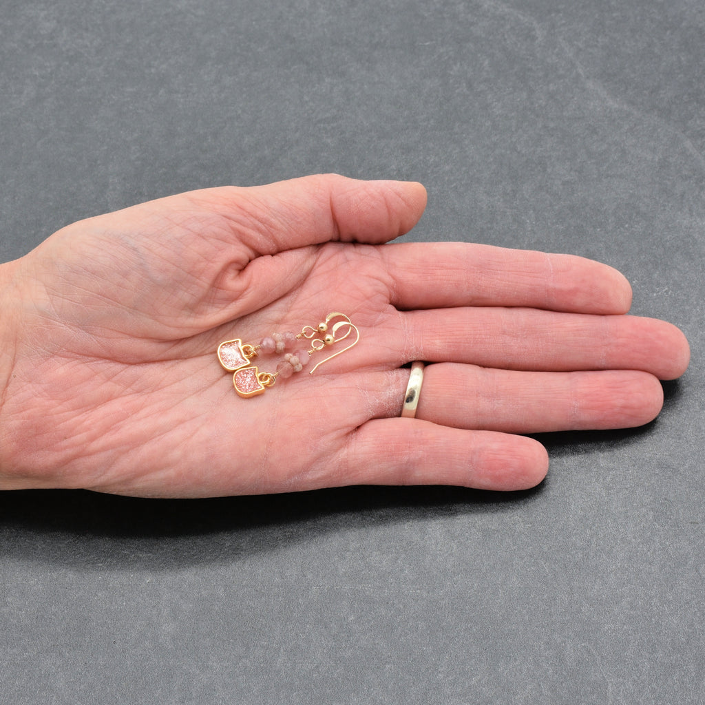 Artist's hand holding lovely delicate pink dangle earrings with cat charms, pink tourmaline, and gold ear wires on a gray background - handmade with love by Ruby Acorn