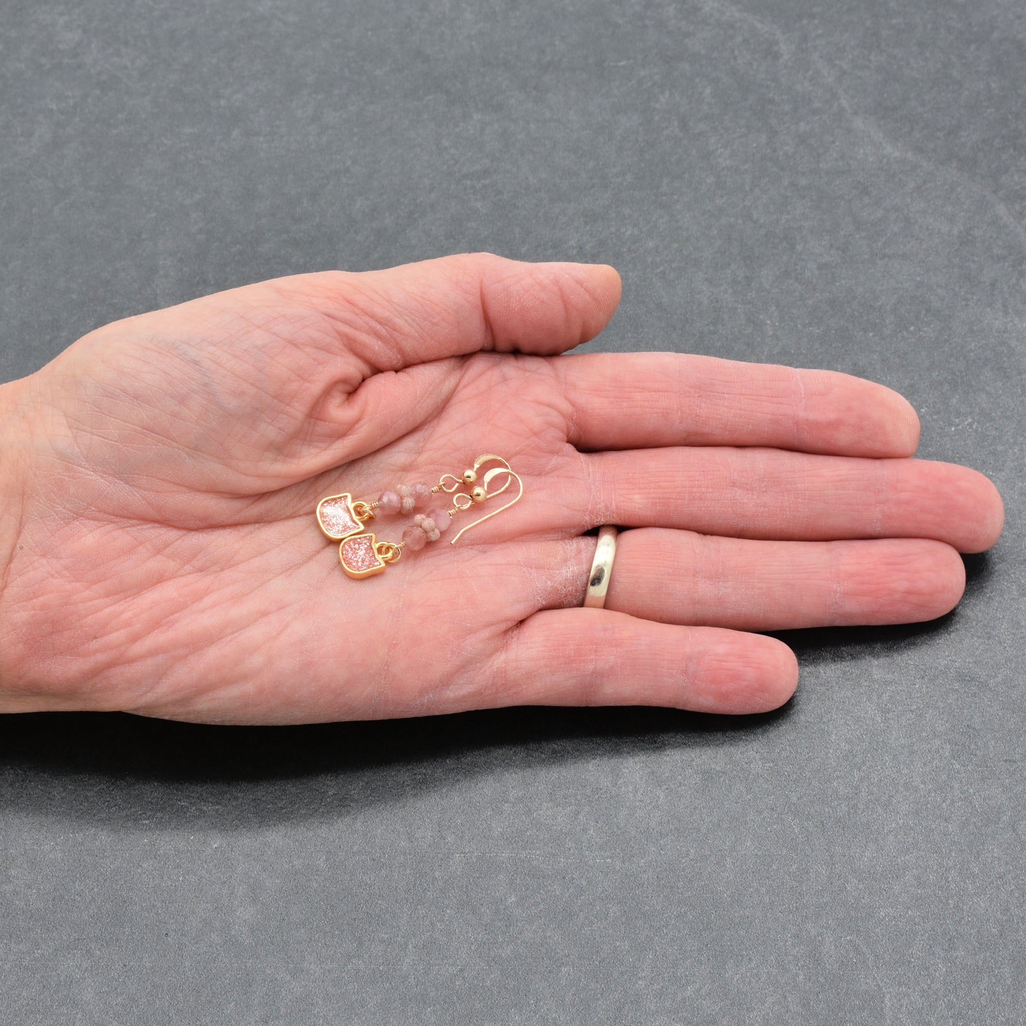 Artist's hand holding lovely delicate pink dangle earrings with cat charms, pink tourmaline, and gold ear wires on a gray background - handmade with love by Ruby Acorn
