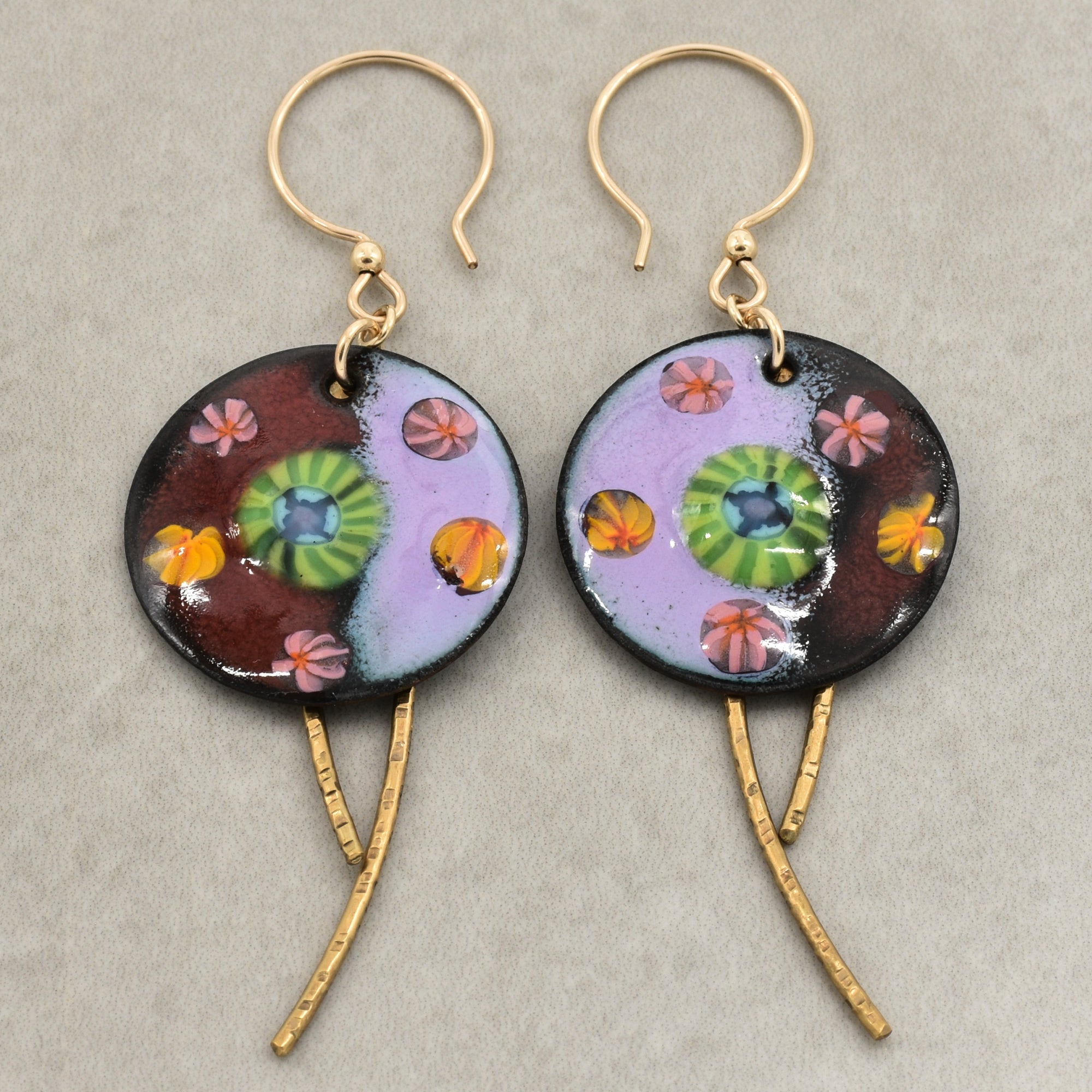 A pair of handmade enamel earring pendants with gold-tone ear wires and brass textured daggers, featuring a colorful Millefiori pattern on a light background - made with love in Colorado by Ruby Acorn.