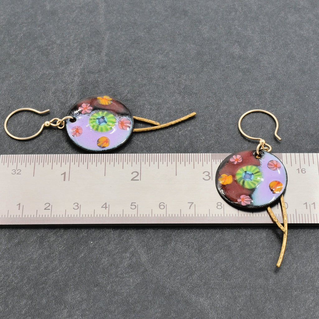 A pair of handmade earrings with gold-tone ear wires, displayed on a dark gray textured background with a metal ruler for scale. The enamel charms feature a colorful mottled pattern of lavender, burgundy, green, and orange - made with love in Colorado by Ruby Acorn.