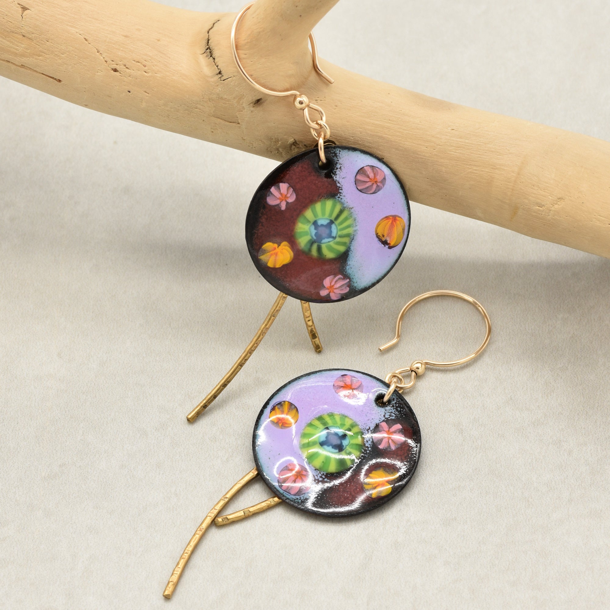 A pair of handmade circular drop earrings with gold fishhook wires and gold branch-shaped dangling elements. The round top pieces feature a multicolor enamel pattern with purple, green, orange, and pink details on a dark background - made with love in Colorado by Ruby Acorn.