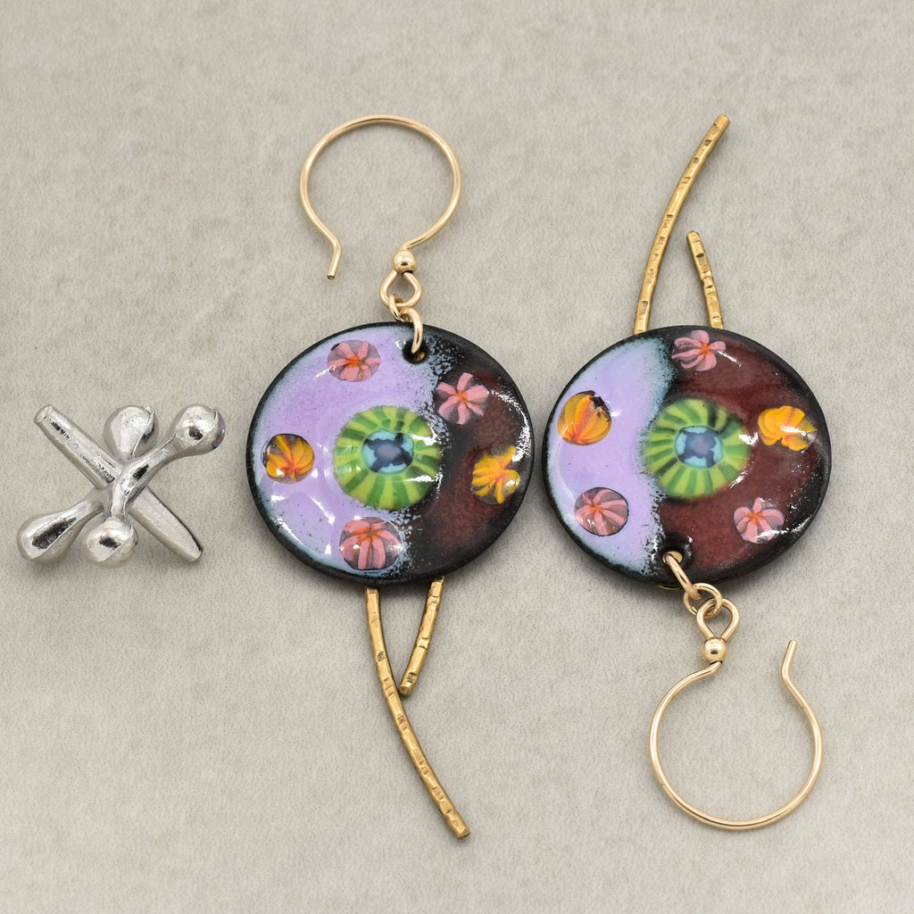 A pair of handmade circular dangle earrings with gold ear wires and brass daggers, featuring colorful artisan enamel art charms with purple, green, orange, and pink floral motifs. A small silver flower-shaped charm is placed beside them on a neutral gray background - made with love in Colorado by Ruby Acorn.