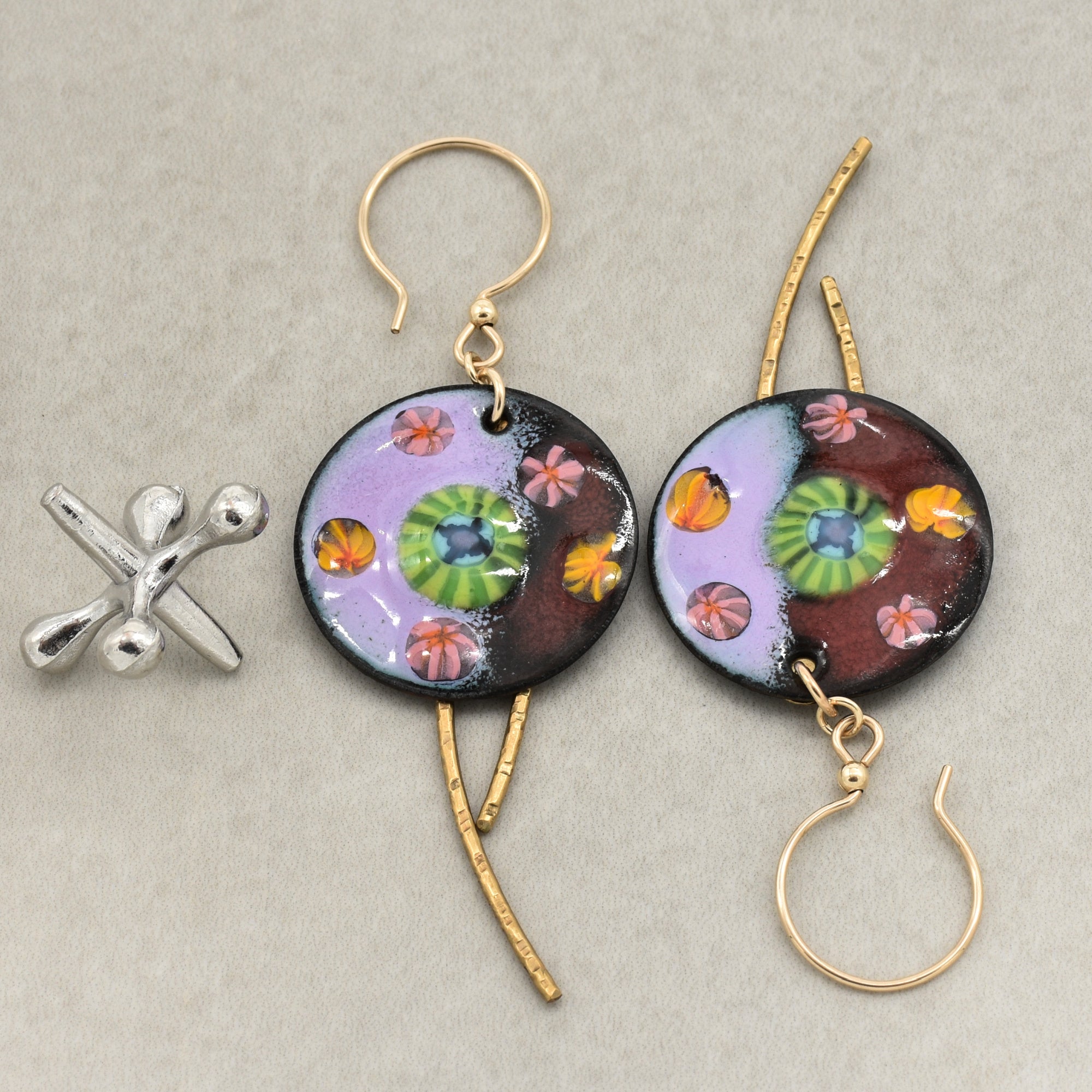 A pair of handmade circular dangle earrings with gold ear wires and brass daggers, featuring colorful artisan enamel art charms with purple, green, orange, and pink floral motifs. A small silver flower-shaped charm is placed beside them on a neutral gray background - made with love in Colorado by Ruby Acorn.