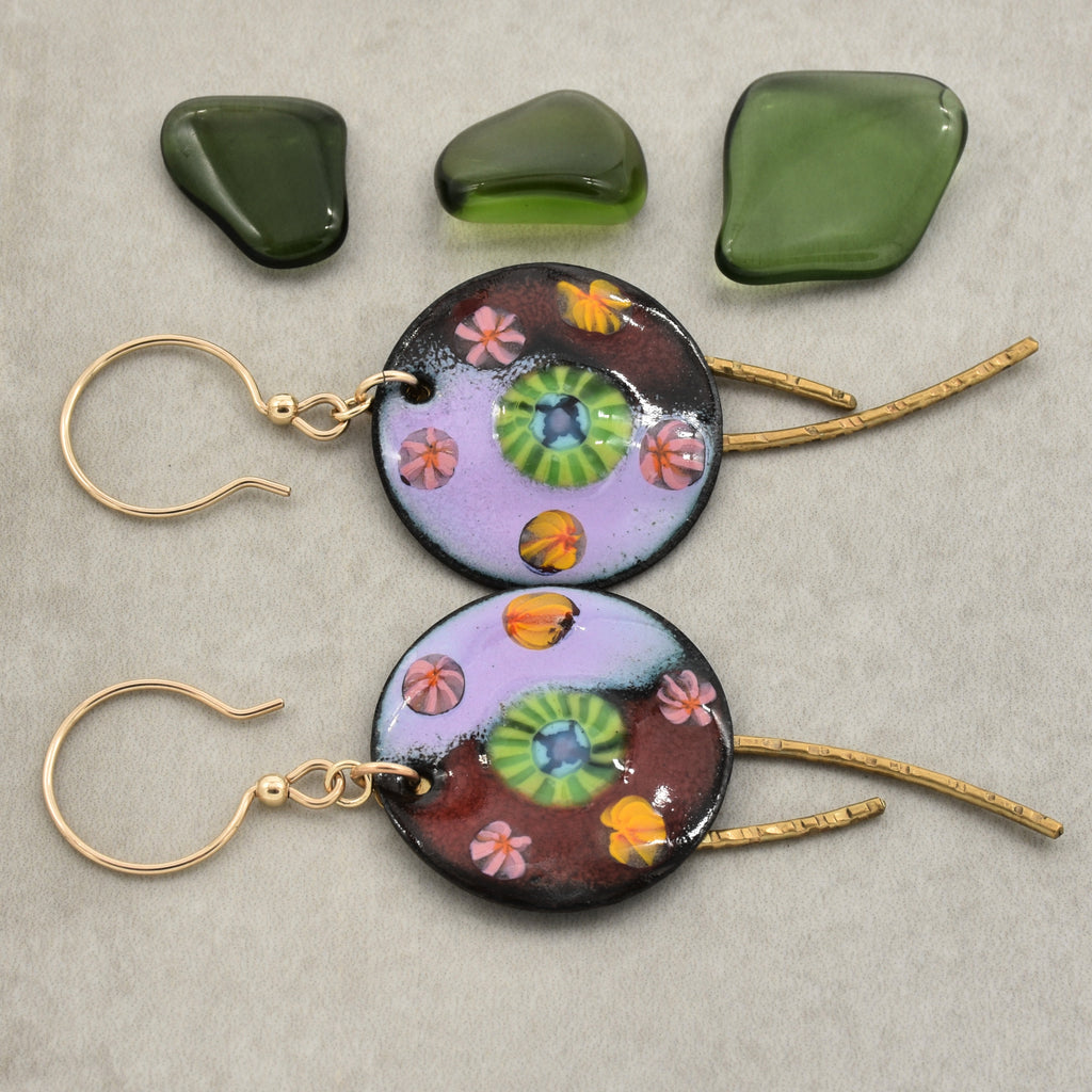 Two handmade round dangle earrings with gold ear wires and textured brass daggers, featuring colorful artisan enamel charms with green, pink, orange, and purple details. Three green polished stones are displayed above the earrings on a neutral gray background - made with love in Colorado by Ruby Acorn.