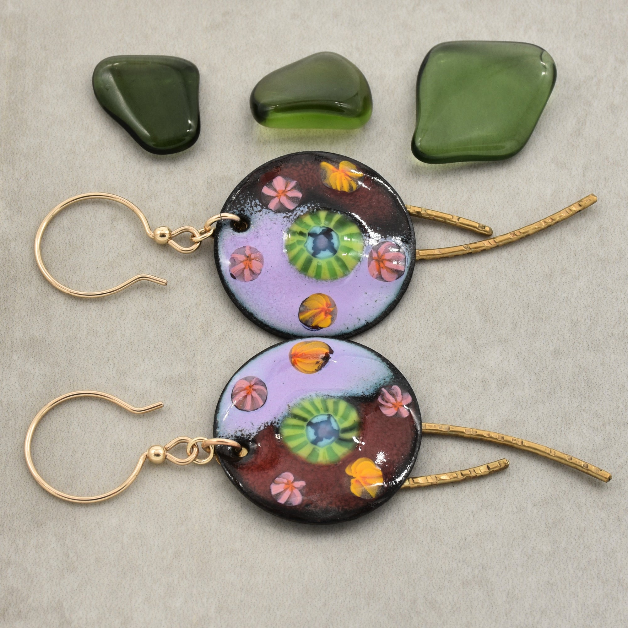 Two handmade round dangle earrings with gold ear wires and textured brass daggers, featuring colorful artisan enamel charms with green, pink, orange, and purple details. Three green polished stones are displayed above the earrings on a neutral gray background - made with love in Colorado by Ruby Acorn.