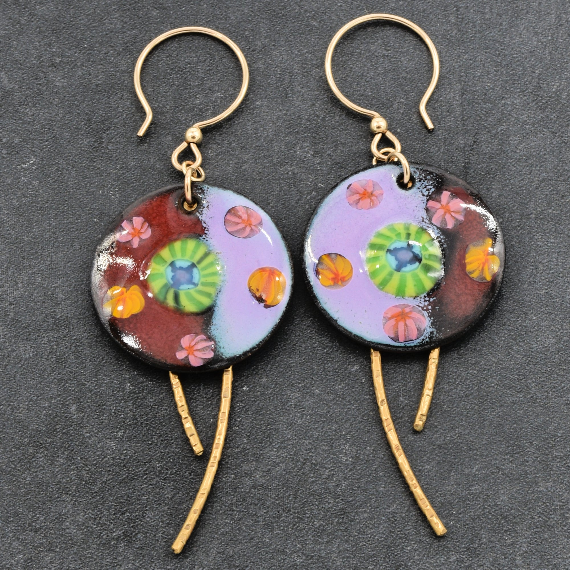 A pair of handmade dangle earrings with circular enamel discs featuring a colorful floral pattern, gold-tone ear wires, and brass daggers, displayed on a dark textured background - made with love in Colorado by Ruby Acorn.