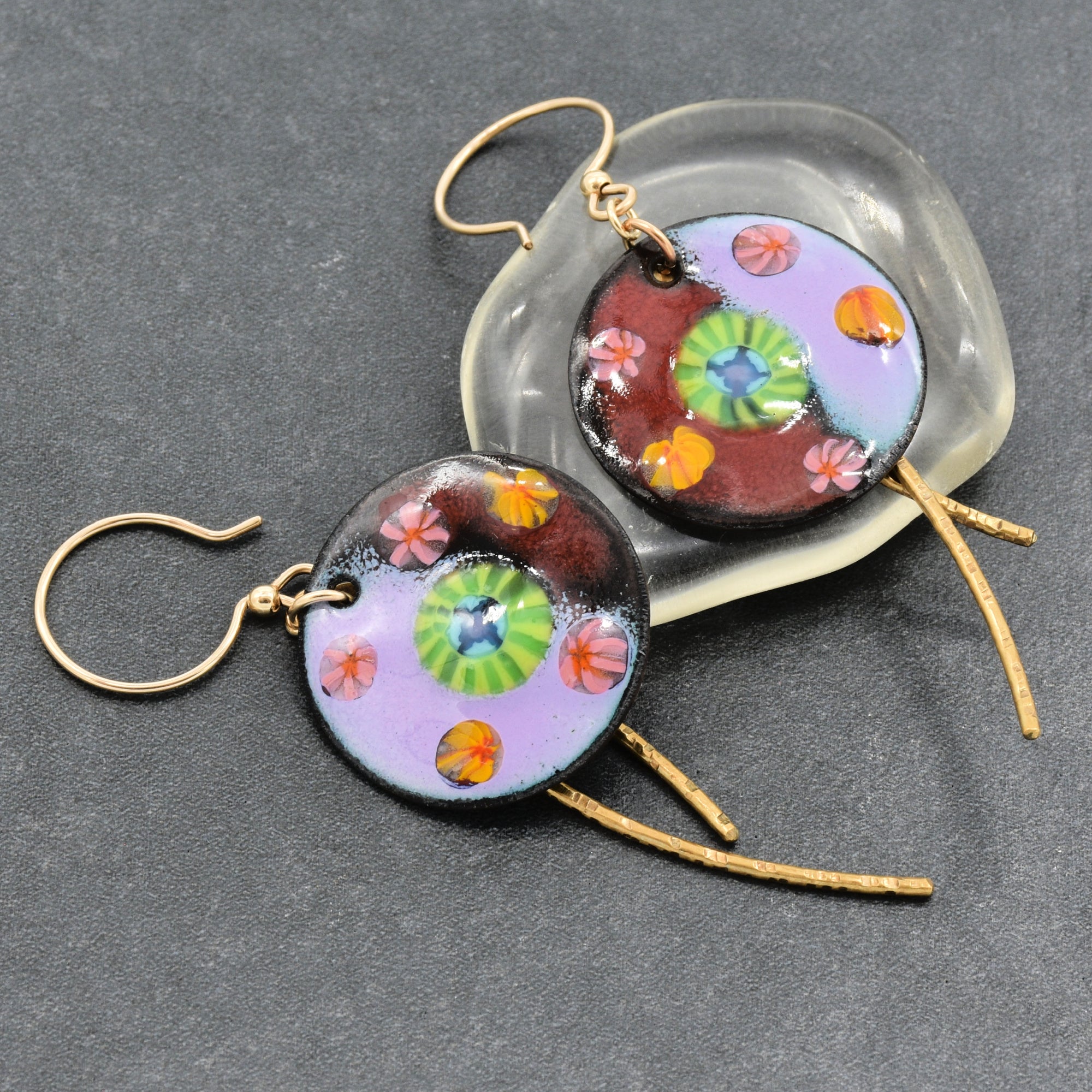 Earrings with artisan enamel  circular charms with gold-tone ear wires and brass daggers, displayed on a dark gray textured background with a clear glass display - made with love in Colorado by Ruby Acorn.