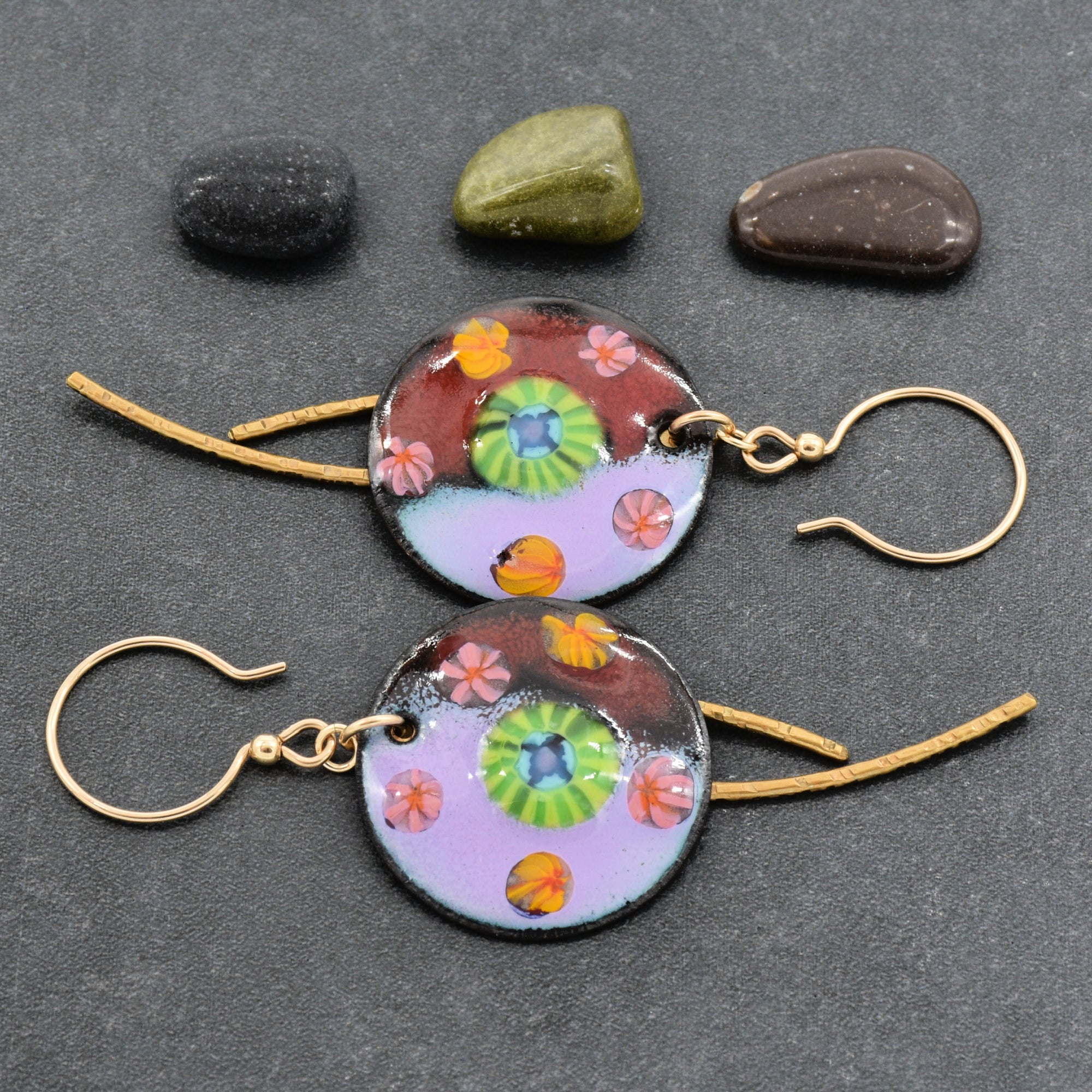 A pair of handmade round enamel charm earrings with gold-tone ear wires and colorful floral motifs, displayed on a dark gray textured background with three small decorative stones - made with love in Colorado by Ruby Acorn.