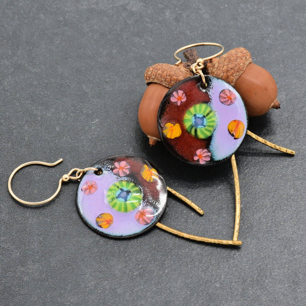 A pair of handmade dangle earrings with round enamel disc charms featuring a colorful floral and geometric pattern, suspended from gold-tone wire and hooks, displayed on a dark gray textured background - made with love in Colorado by Ruby Acorn.